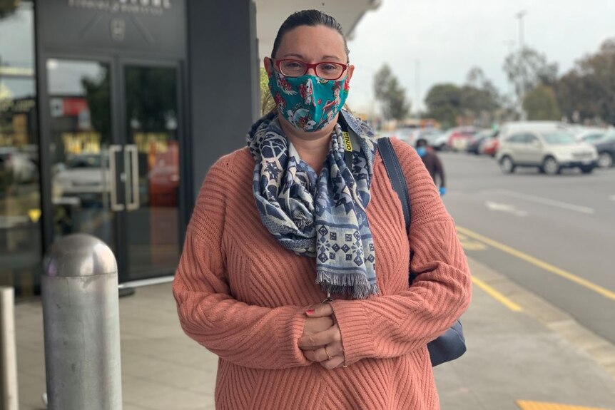 A woman with red glasses, a green face mask and peach jumper stands in front of a shopping centre.