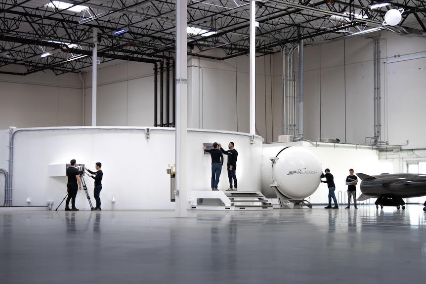 People in a wide space, concrete floors, white round walls or tanks, a black rocket on stand.