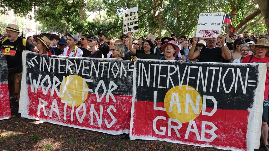 A group of people standing with Aboriginal flag banners that read "intervention = land grab'.
