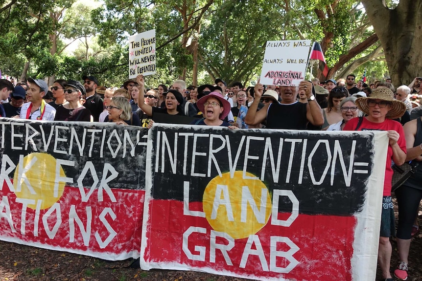A group of people standing with Aboriginal flag banners that read "intervention = land grab'.