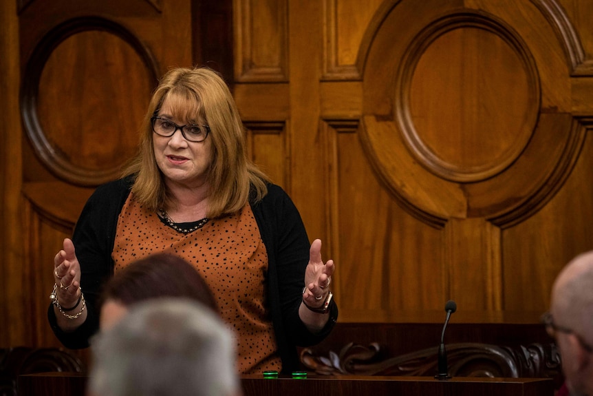 A woman with glasses and red hair gesticulates in a wood-panelled room