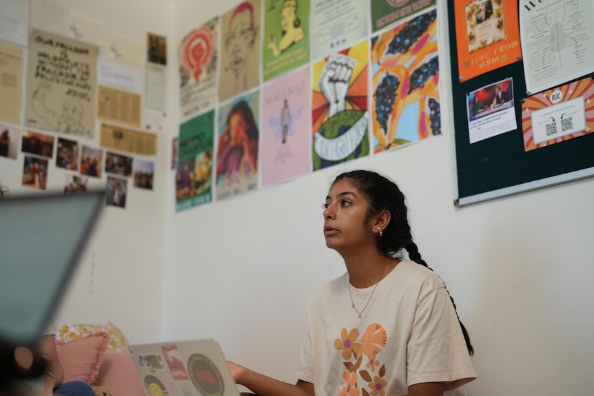 A young woman with long braids sits on a bed. Her wall behind her is covered in posters and memory notes