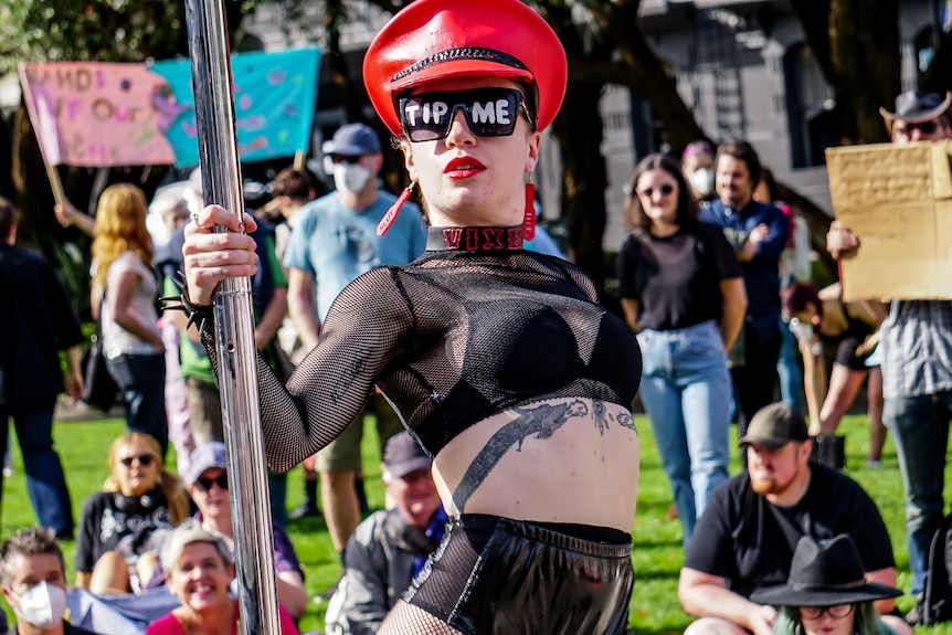 A woman stands next to a stripper pole with the words 'tip me' written on her sunglass lenses 