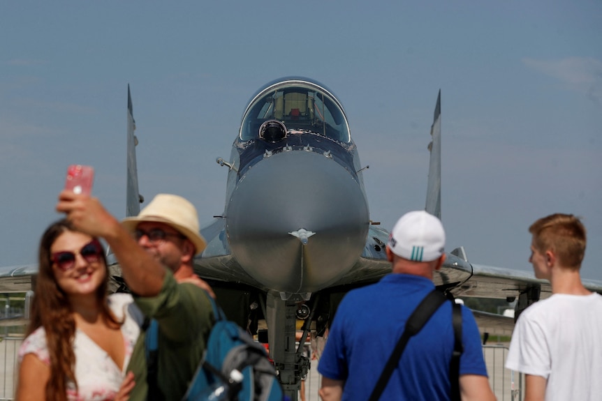 Tourist-looking people pose for pictures in front of a fighter jet.