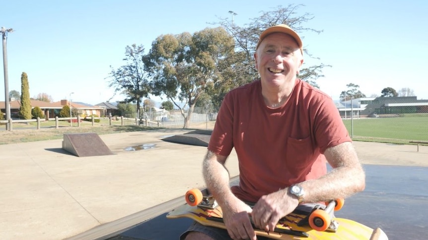 A man in a red shirt and wearing a cap holds an orange skateboard upside down while sitting on a skate ramp 