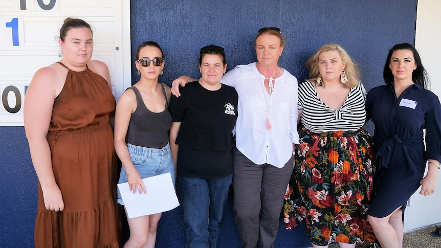 A group of six women all looking upset standing in front of a blue wall.