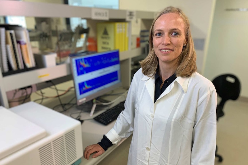 A woman with blonde hair stands in a lab in front of a computer wearing a lab coat and posing for a photo.