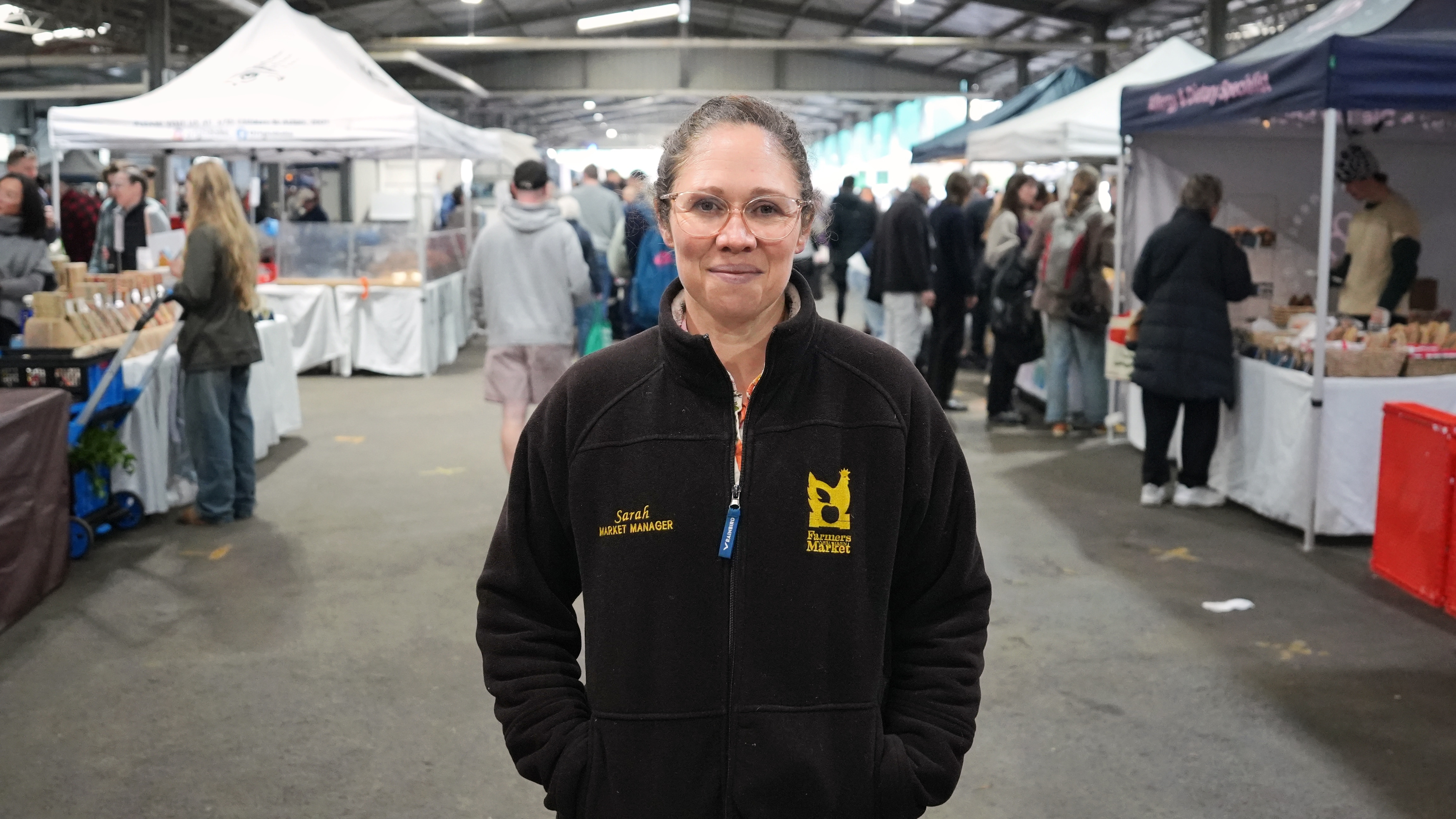 A woman with light hair pulled into a ponytail stands in a warehouse surrounded by market stalls.