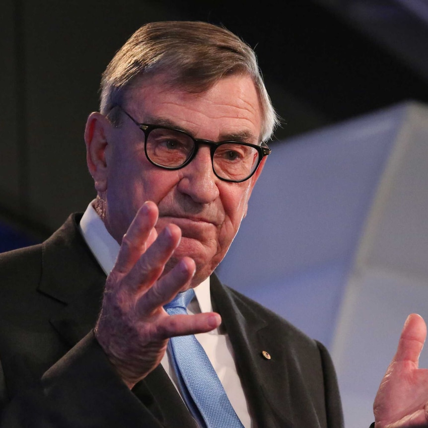 John Lord stands at the podium at the National Press Club, gesticulating with his hands and wearing thick rimmed glasses.