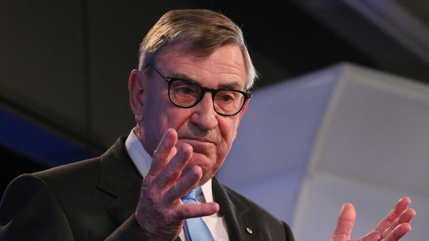 John Lord stands at the podium at the National Press Club, gesticulating with his hands and wearing thick rimmed glasses.