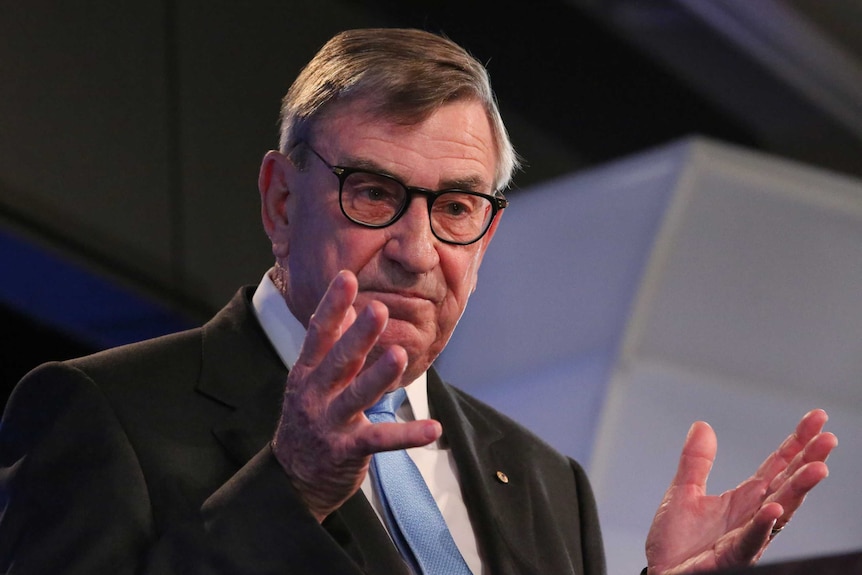 John Lord stands at the podium at the National Press Club, gesticulating with his hands and wearing thick rimmed glasses.