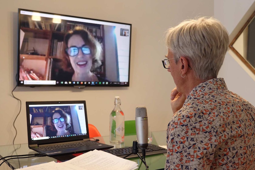 A man has a conversation with a woman via a video conference on a computer which is mirrored onto a large wall mounted screen