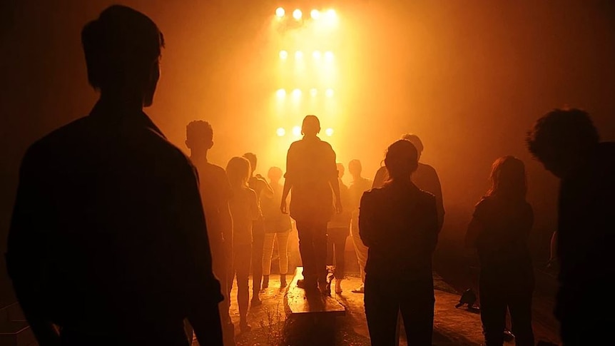The silhouettes of a group of people on a stage, they are facing towards a wall of yellow light