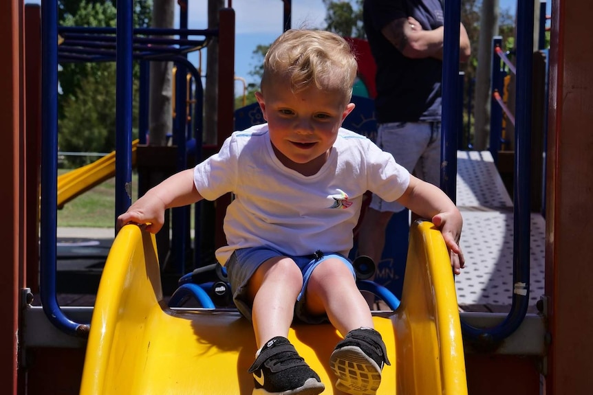 A little boy smiles at the top of a yellow slide