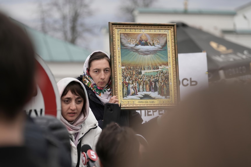Two women in white head scarves, with one woman holding a religious painting.