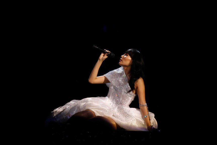 Dami Im dressed in white sitting against a dark backdrop singing into a microphone 