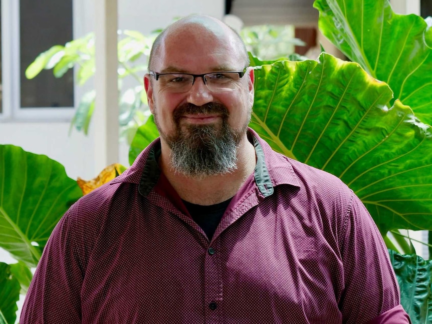 A man in a purple shirt and glass smiling in front of a plant.