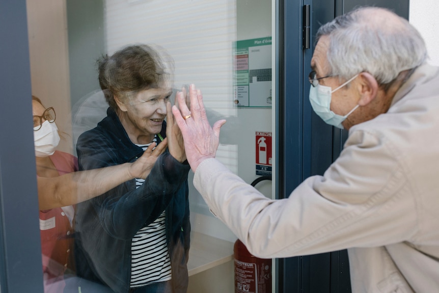 An elderly man and woman hold their hands together through a glass window.