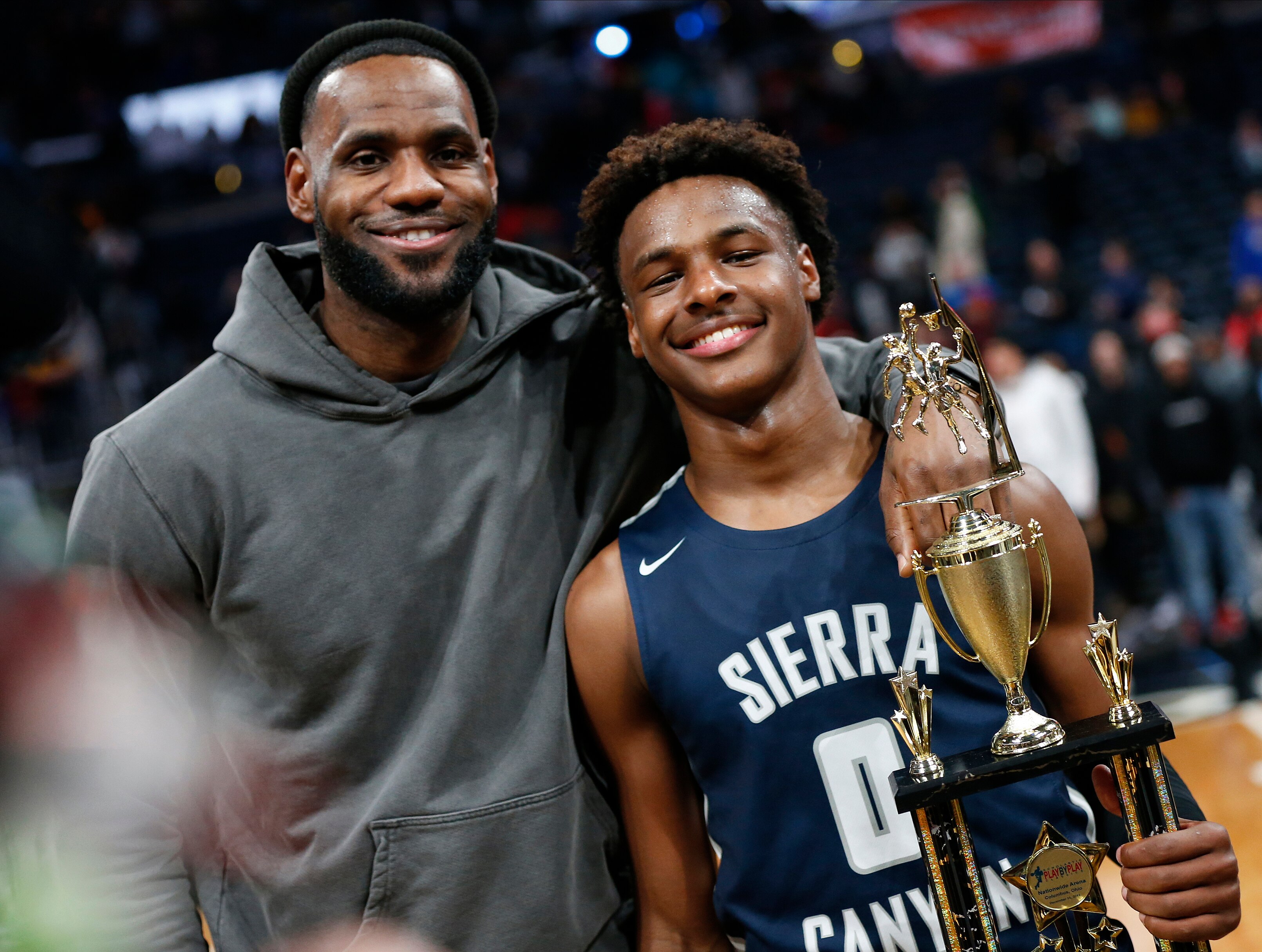 Lebron James with his arm around his son who is holding a trophy and smiling