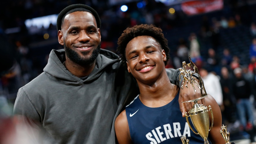 Lebron James with his arm around his son who is holding a trophy and smiling