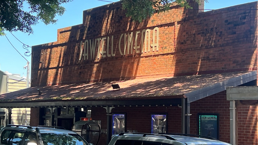 The exterior of an Art Deco brick building with Sawtell cinema written on facade in cream letters, cars parked in front.