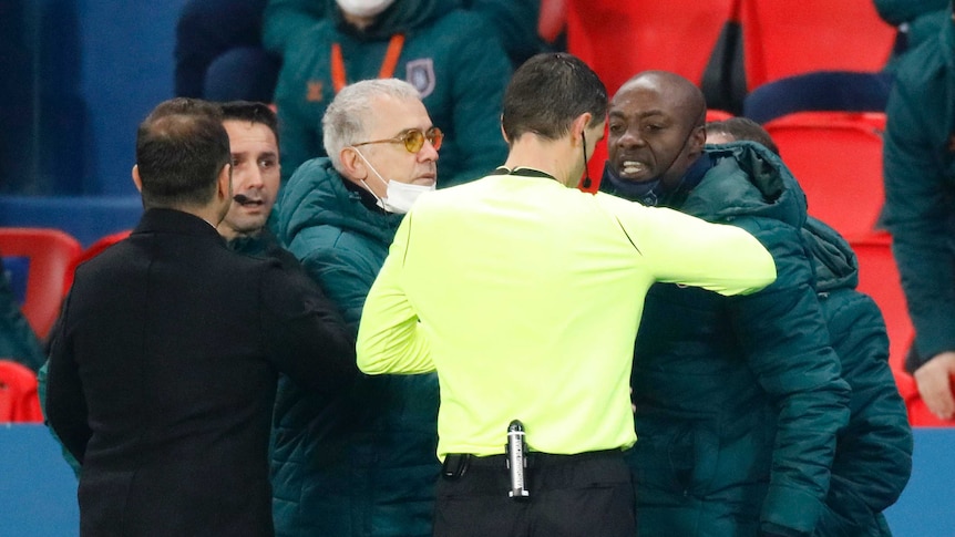 A referee in yellow looks at a man who is grimacing at him, with other people in coats nearby