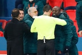 A referee in yellow looks at a man who is grimacing at him, with other people in coats nearby