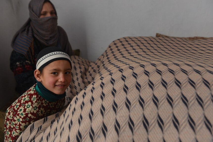 A little boy smiles at the camera, huddled under a large blanket. His mother sits behind, watching