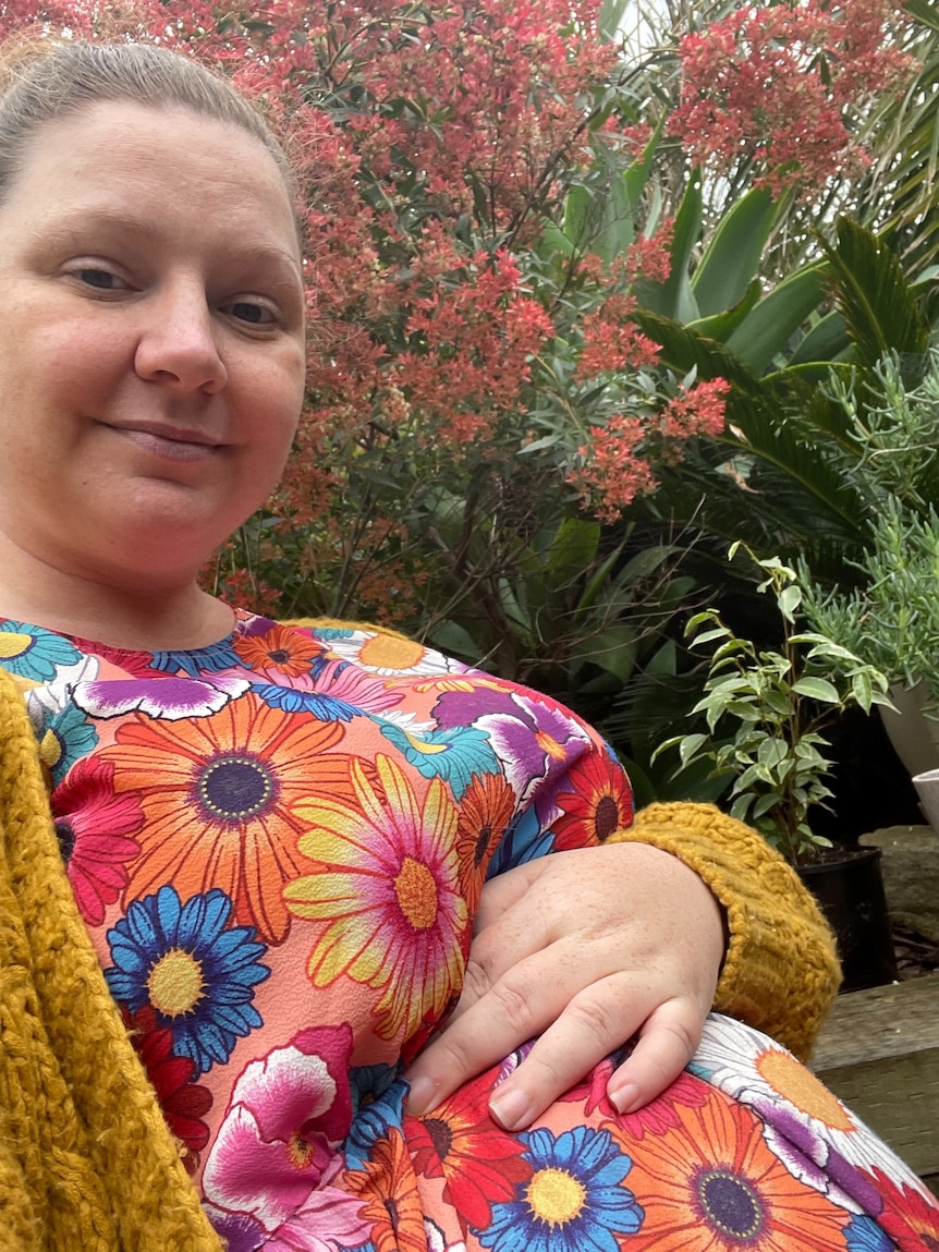 A woman in a colourful floral dress and yellow cardi nurses her baby belly.