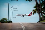 A QantasLink plane flies over a street between a row of streetlights and palm trees.