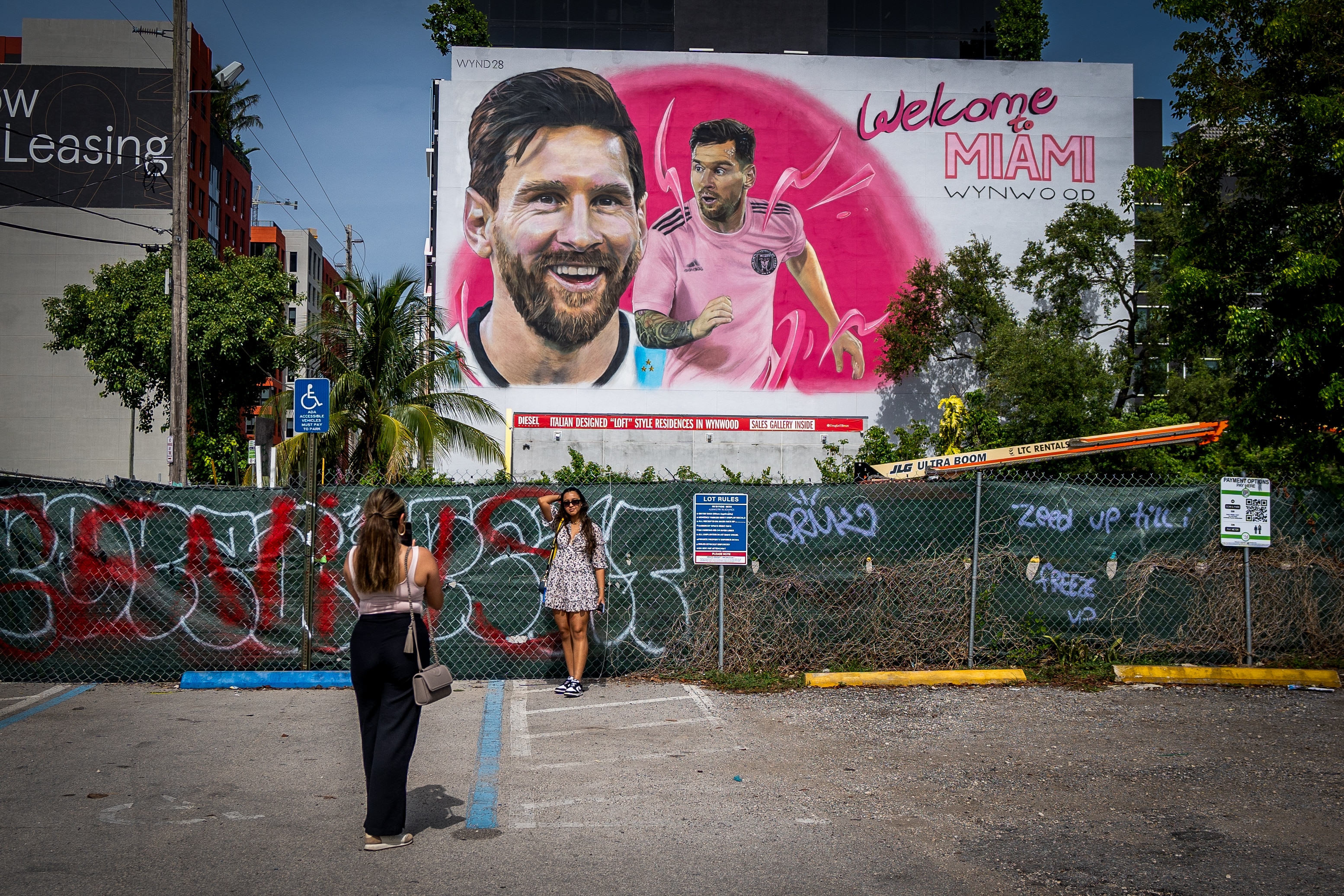 A woman poses as her friend takes a picture of her with a giant mural titled WELCOME TO MIAMI with Messi's face