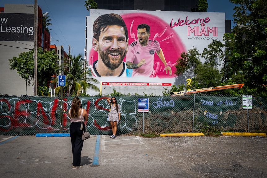 A woman poses as her friend takes a picture of her with a giant mural titled WELCOME TO MIAMI with Messi's face