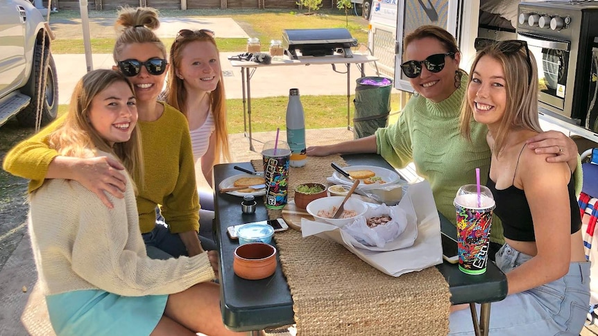 Five women sit around a table outside a caravan