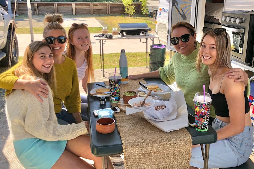 Five women sit around a table outside a caravan