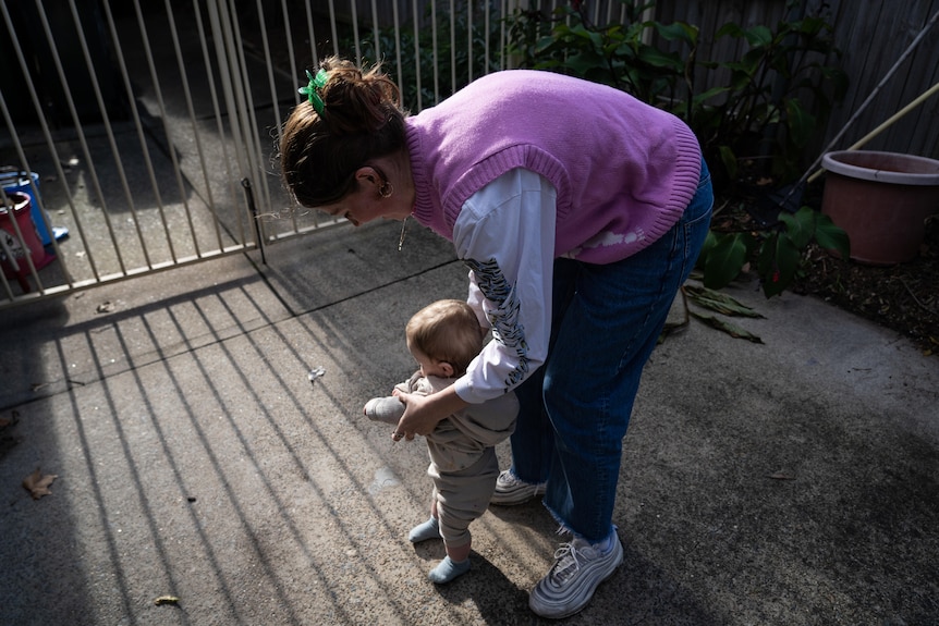 A young woman crouches down and plays with a baby in a concrete courtyard.