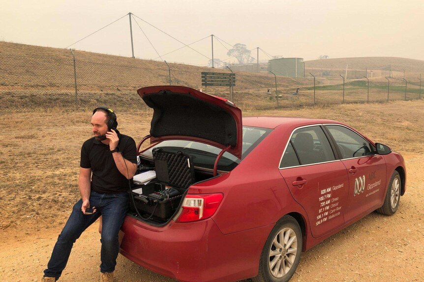 Kendall with headphones on sitting on bumper of ABC car with broadcast equipment in the boot amid smoky surroundings on a hill.