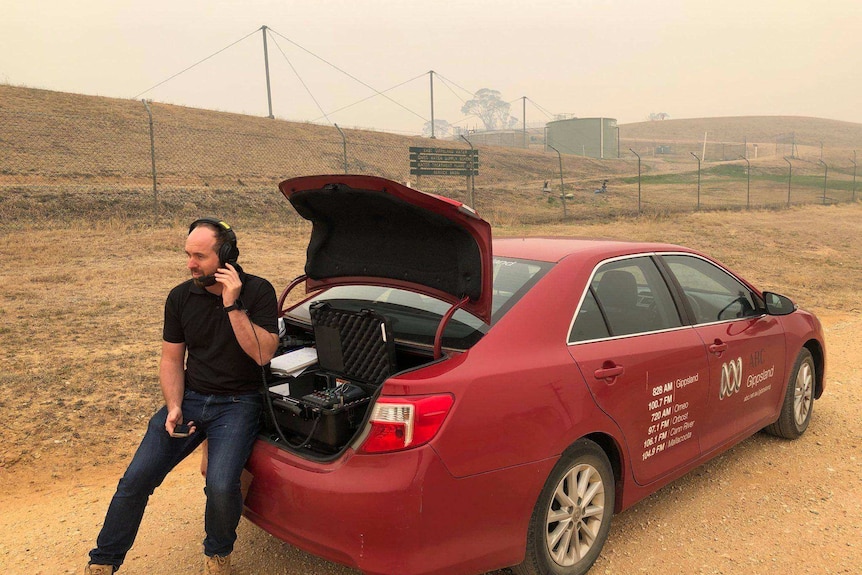 Kendall with headphones on sitting on bumper of ABC car with broadcast equipment in the boot amid smoky surroundings on a hill.