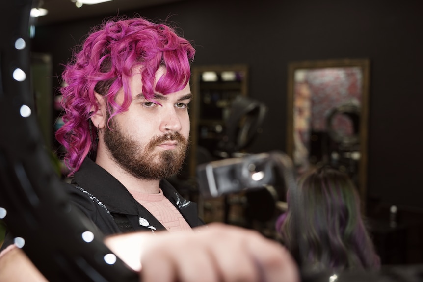 A hairdresser with bright pink hair sets up a ring light, about to photograph his client.