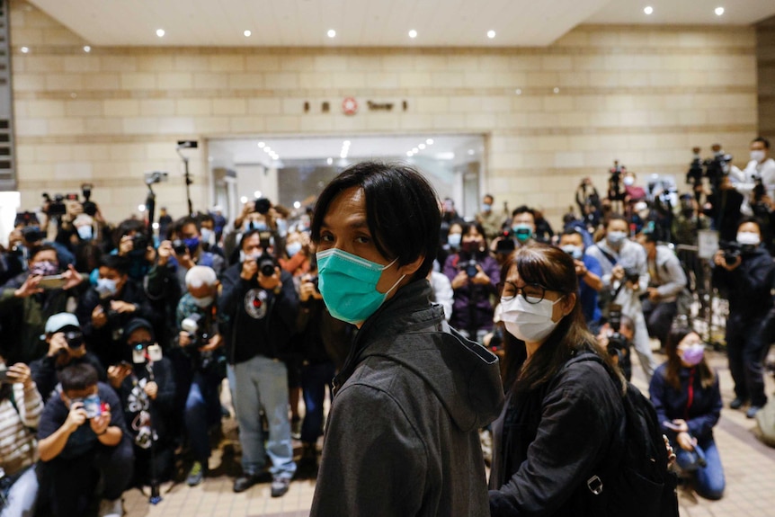 A young man in a green mask walks hand-in-hand with woman as crowd of photographers surround them.