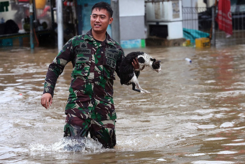 A man smiles in army fatigues and holds a damp black and white cat in one hand as he wades through muddy water in floods.
