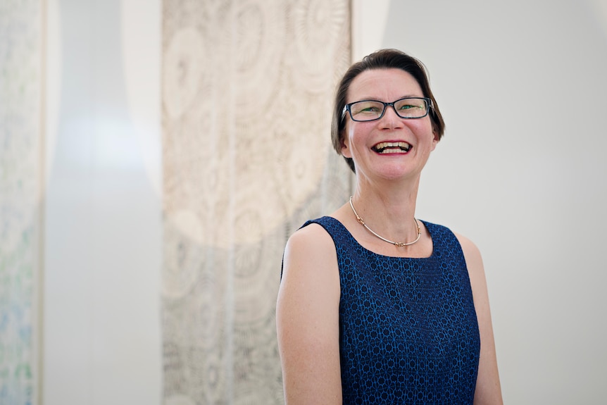 A middle-aged white woman wearing a blue dress and glasses