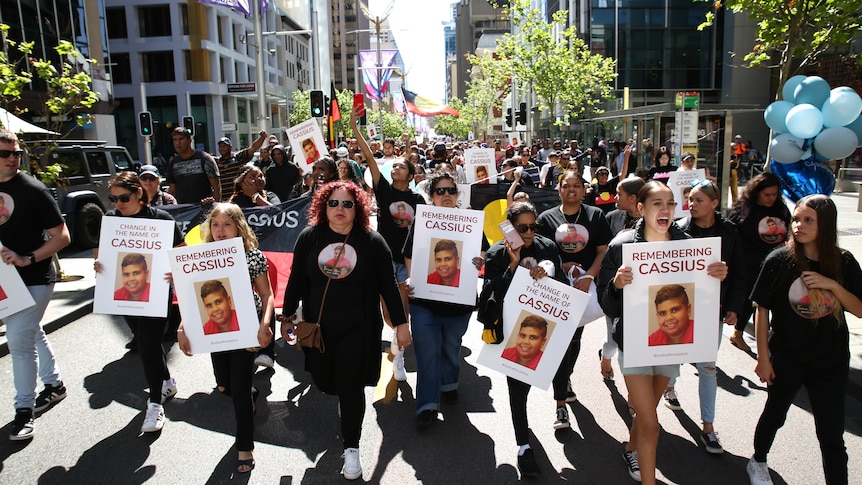 A large group of people match down a city street holding signs saying 'justice for Cassius'
