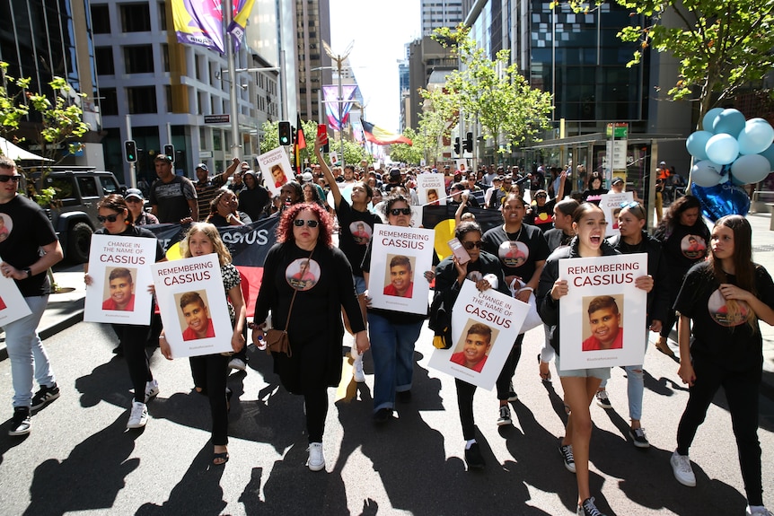 A large group of people match down a city street holding signs saying 'justice for Cassius'