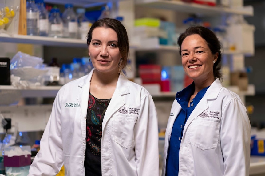 The two scientists stand in a lab wearing white lab coats and smiling happily.