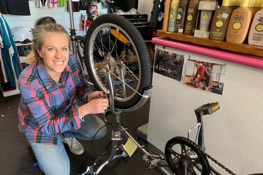 A smiling woman kneeling down and tinkering with a bicycle.