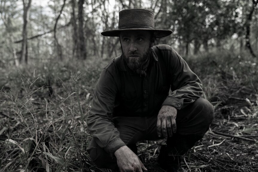 A desaturated image of a man in a hat, wearing 19th-century dress, with a determined expression, crouching in undergrowth.