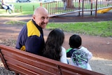 Neil Reynolds looks at the camera over his shoulder on a park bench with the backs of two small children on the bench with him.