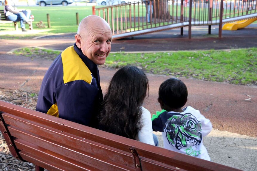 Neil Reynolds looks at the camera over his shoulder on a park bench with the backs of two small children on the bench with him.
