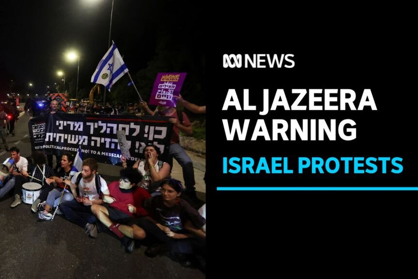 Al Jazeera Warning, Israel Protests: Protesters sitting on road at night in front of banner in Hebrew and Israeli flag.