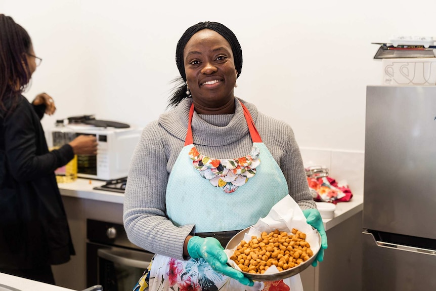 A mid shot of a smiling woman with dark hair standing in a kitchen wearing an apron and holding a bowl of food.